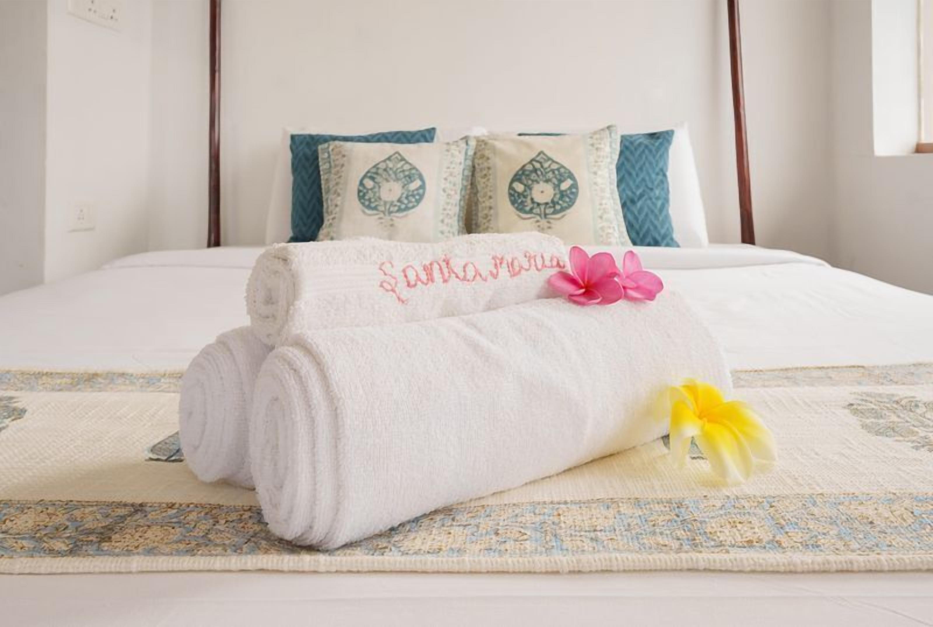 A neatly made bed with a rolled towel and a pink flower on a patterned rug.