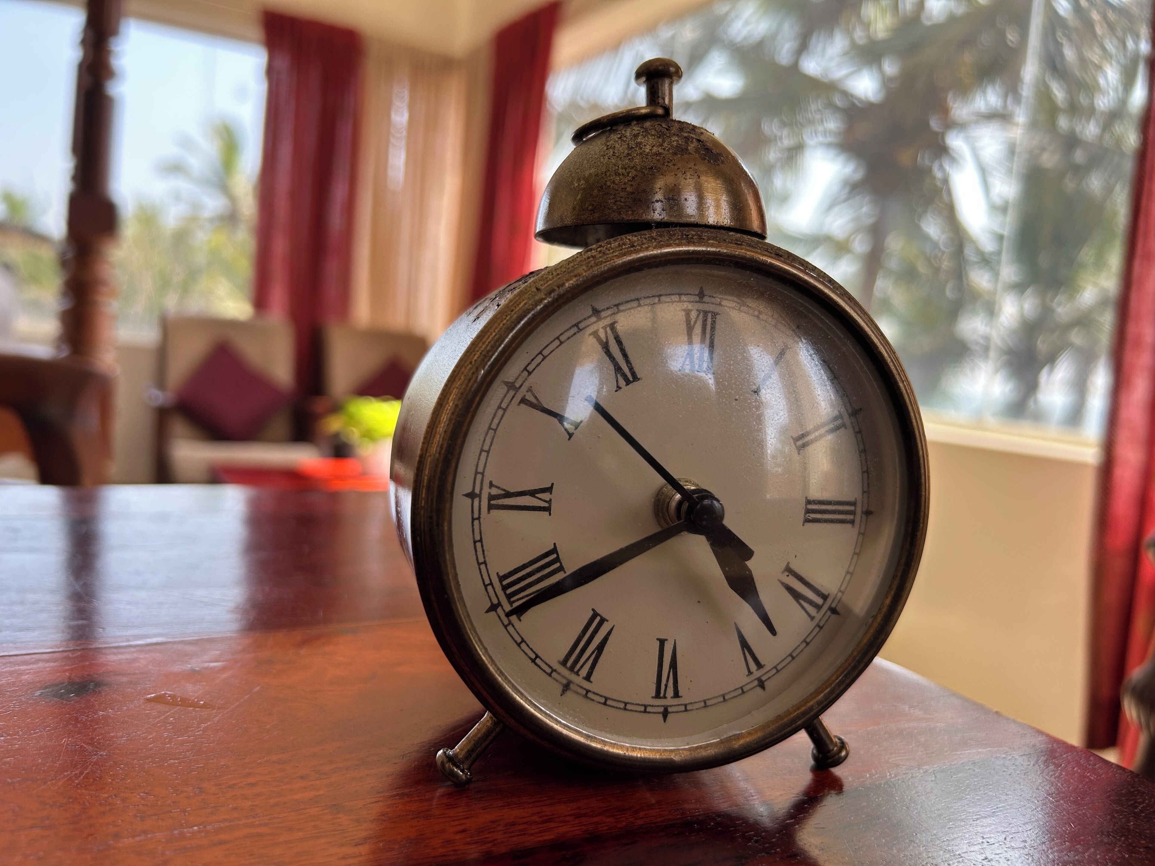 A vintage alarm clock on a wooden table in a room with a view of palm trees.