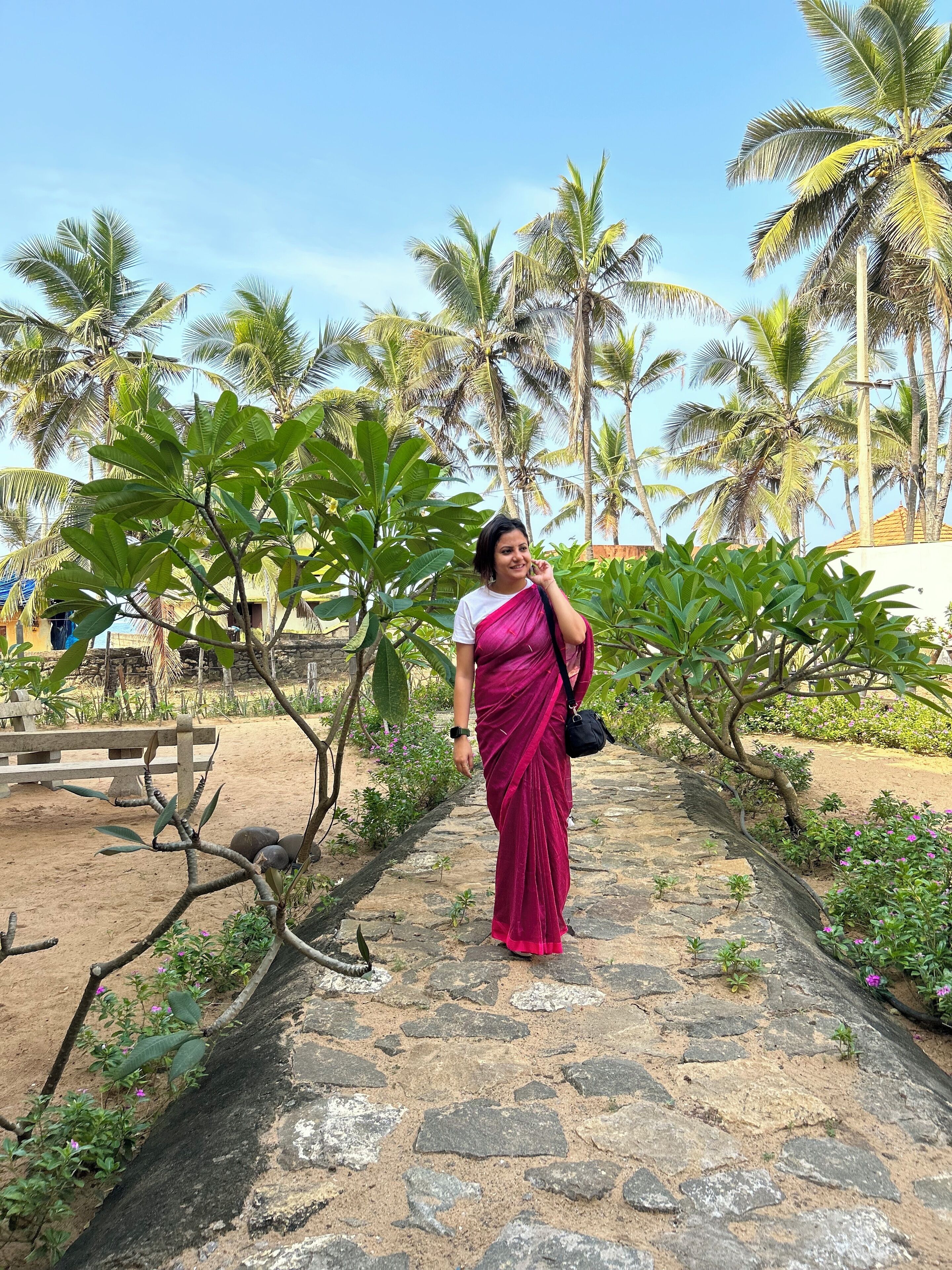 A woman in a pink saree walking on a stone pathway surrounded by greenery and palm trees.