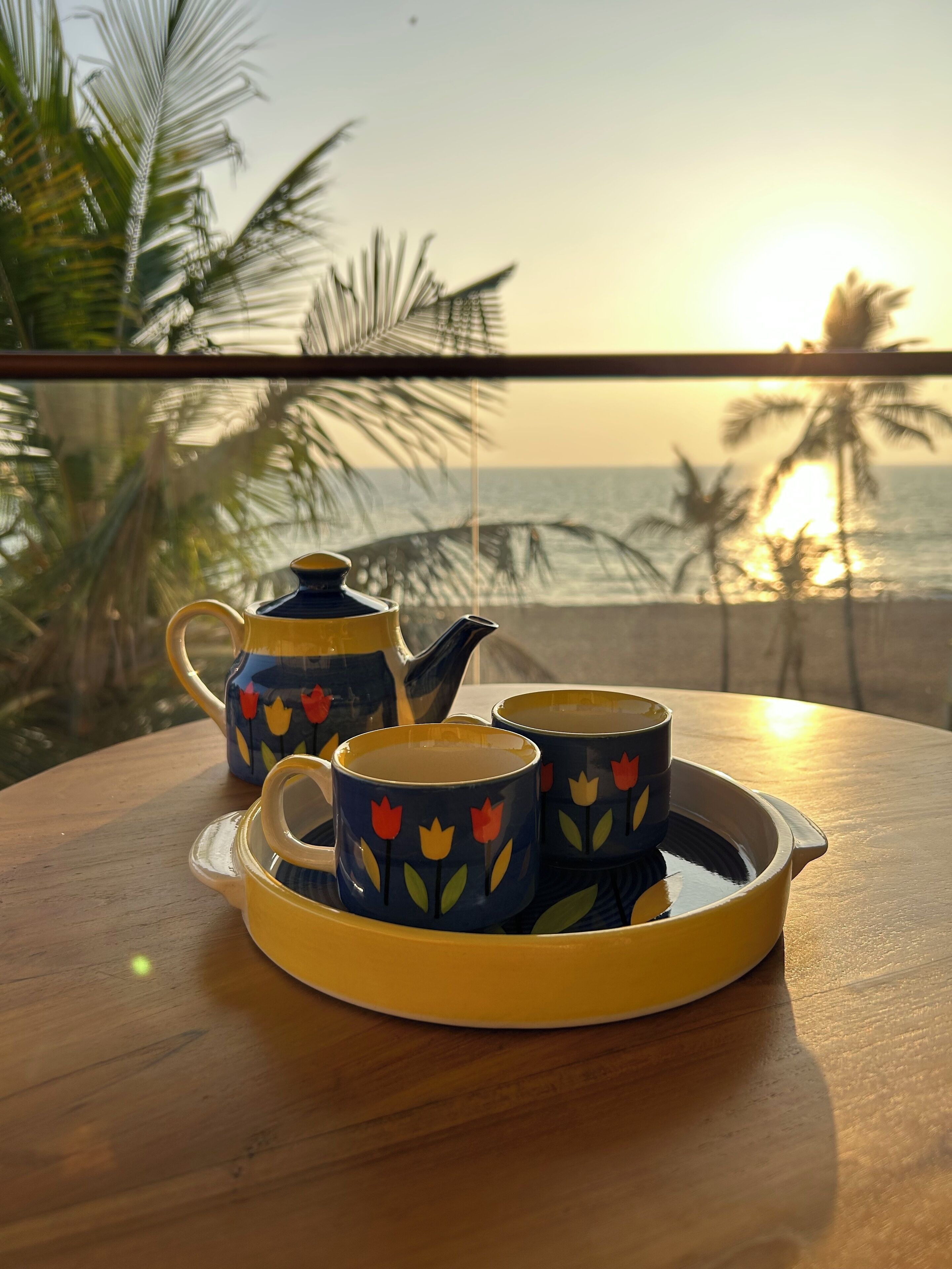 A table with a teapot and two cups, set against a view of the ocean and palm trees at sunset.
