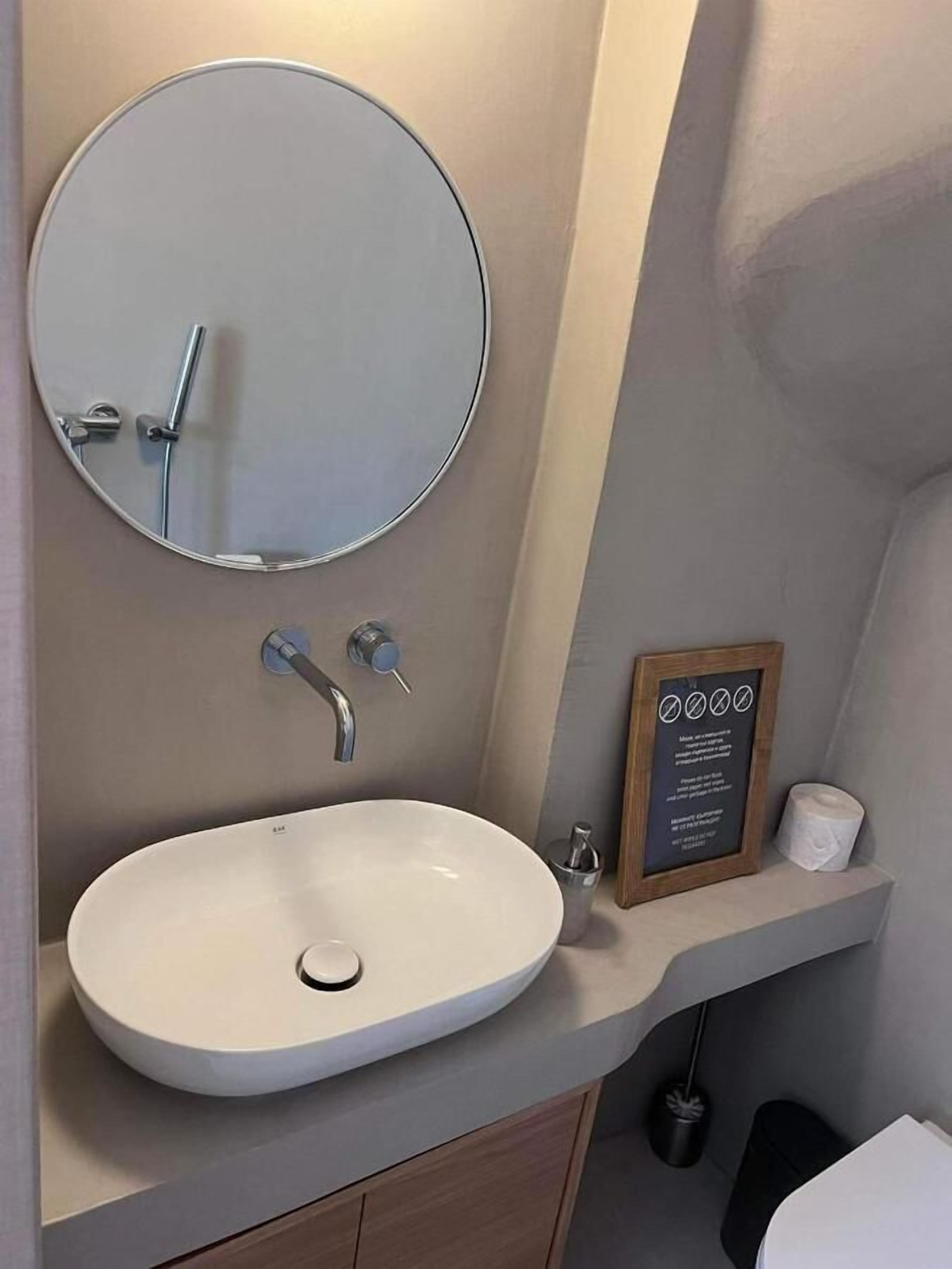Ein Badezimmer mit rundem Spiegel, Waschbecken, einem Schild mit Informationen und Toilette.