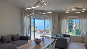 Una sala de estar moderna con una mesa de comedor de madera, sofás grises y vista al mar a través de amplios ventanales.