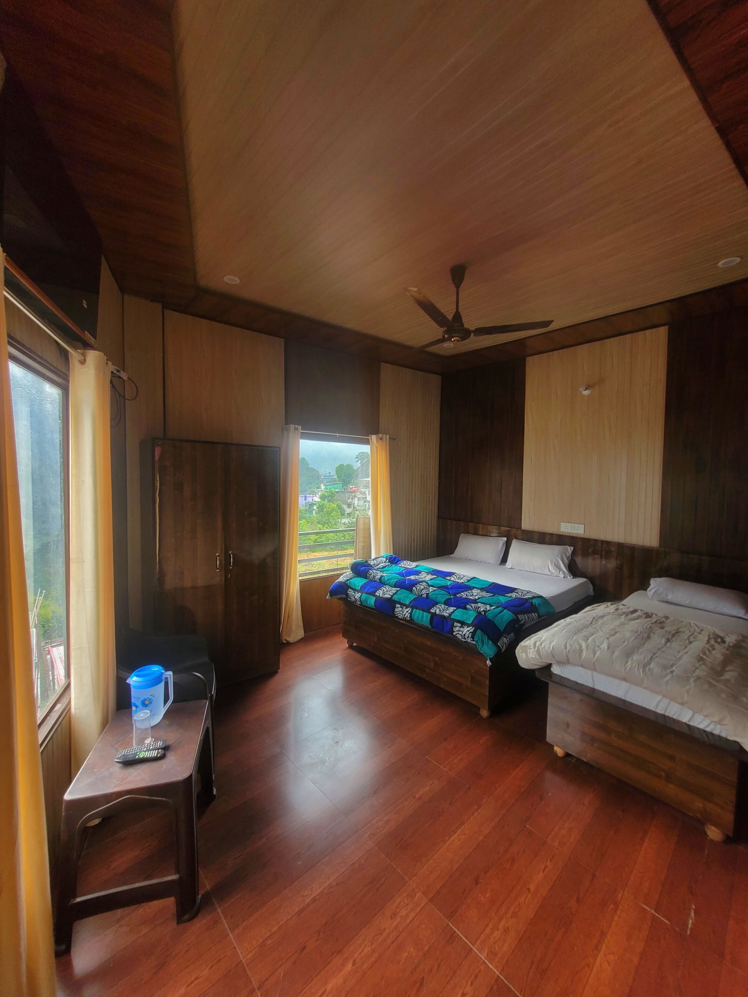 A bedroom with two beds, wooden flooring, a desk with a chair, a ceiling fan, and a window with curtains.