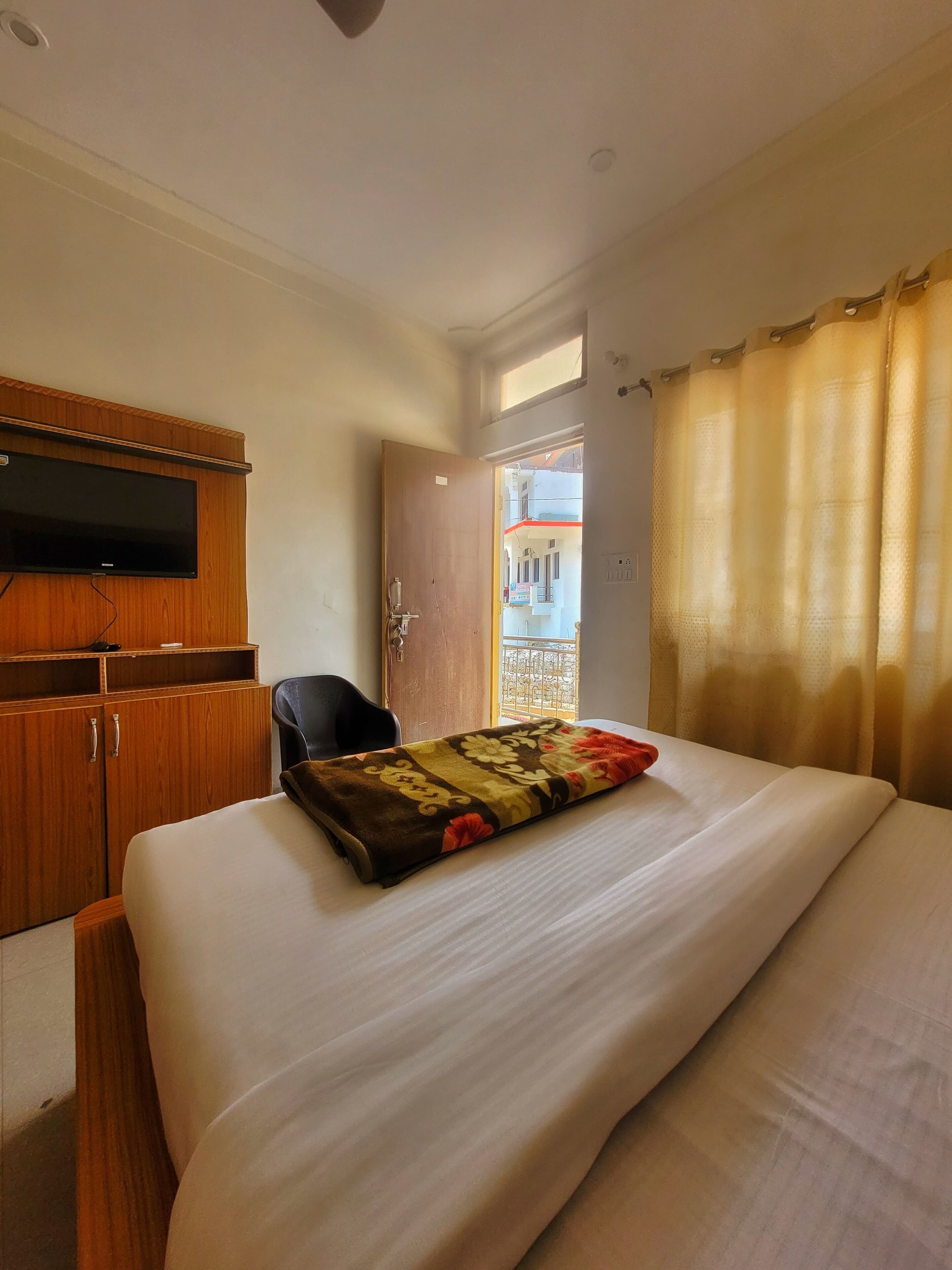 A hotel room with a bed, a wooden wardrobe, a television mounted on the wall, and a window with curtains.