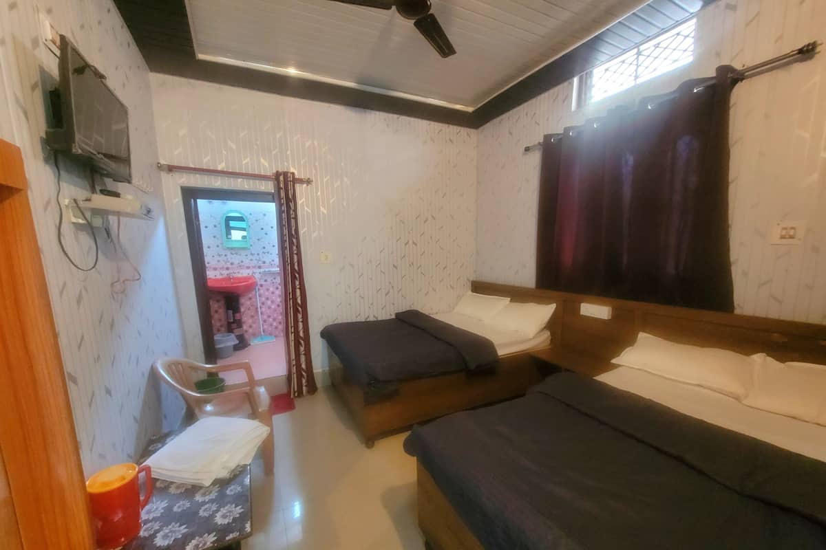 Ein Zimmer mit zwei Betten, einem Schreibtisch, einem Stuhl, einem Fenster mit Vorhängen, einem Deckenventilator und einem kleinen Badezimmer mit Waschbecken.