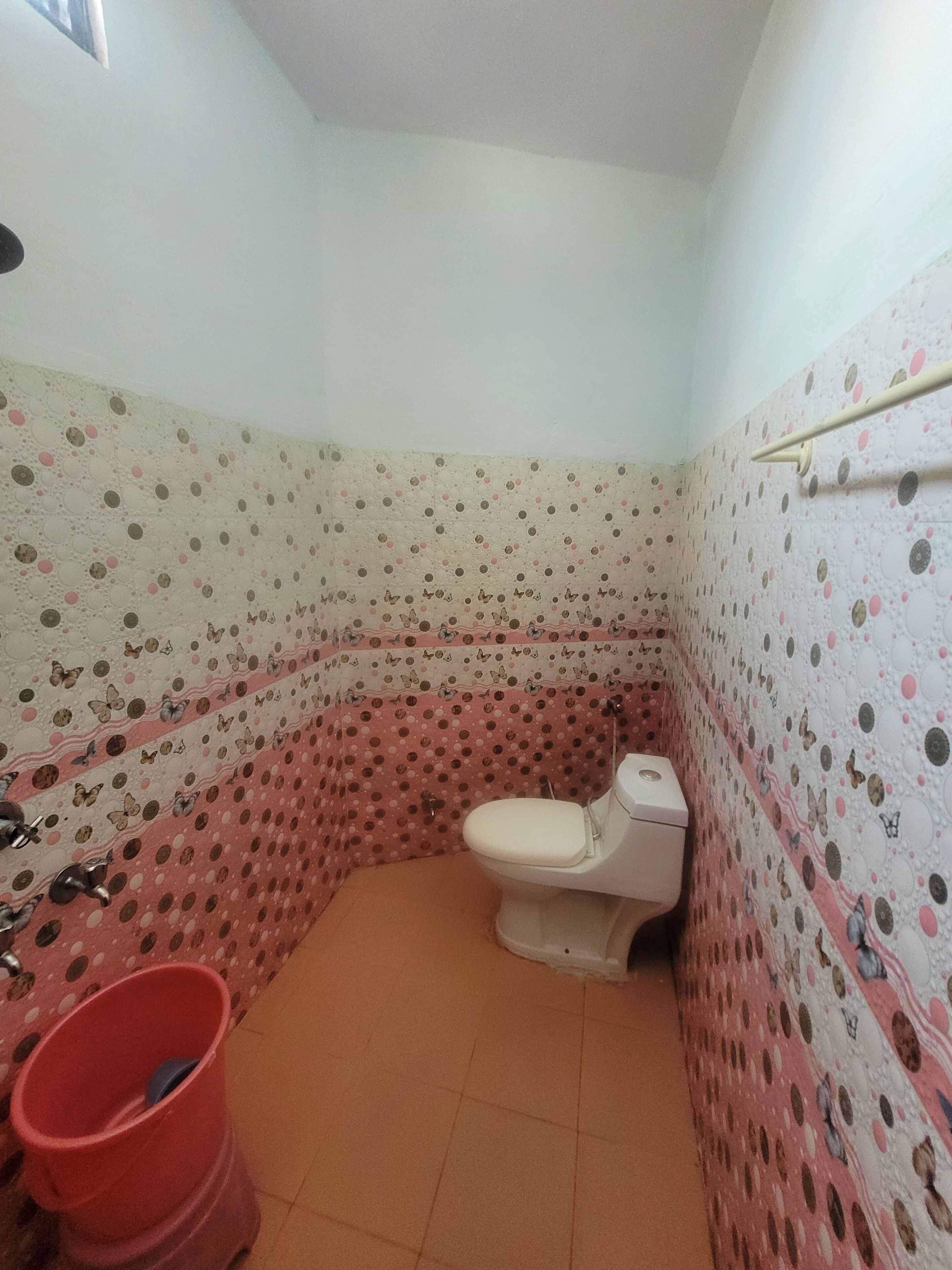 Ein kleines Badezimmer mit Toilette, einem roten Eimer und Wandverkleidung mit Muster.