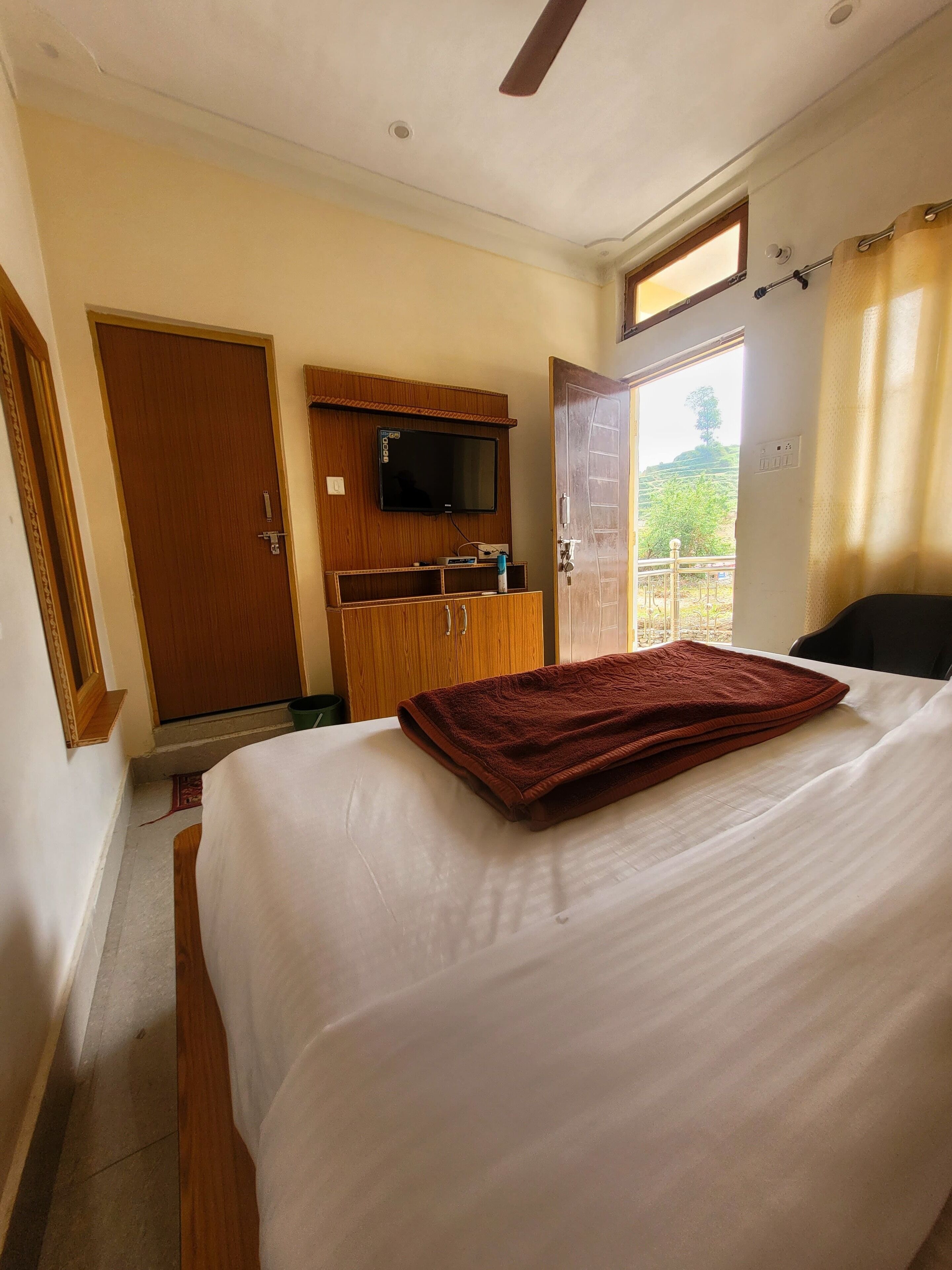 Un dormitorio con cama, televisión sobre una cómoda de madera, ventana con cortinas y una puerta.