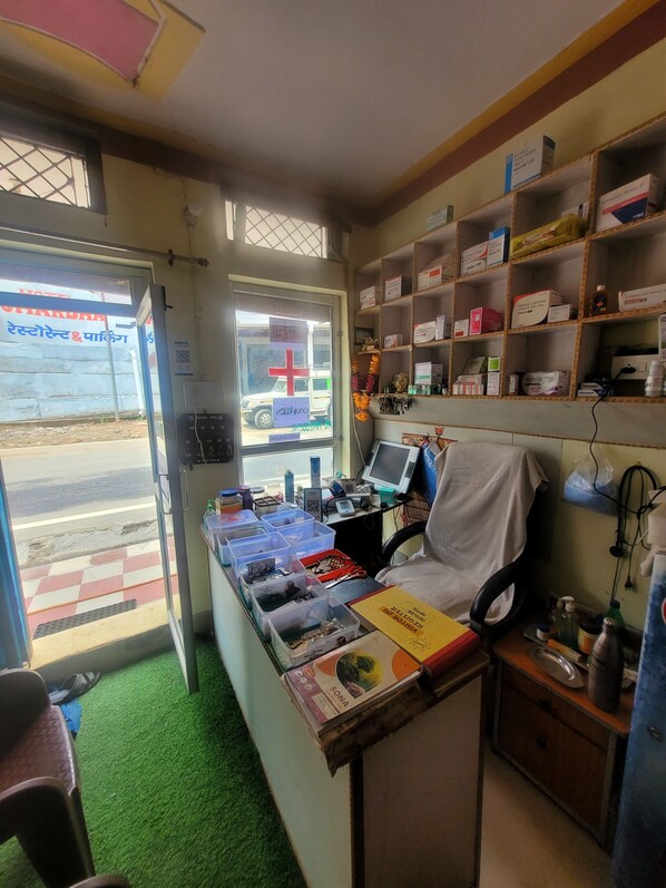 A small, cluttered room with a desk, shelves of medical supplies, a chair, and a window with a view of a street. - Shivlok Hotel (Ukhimath)