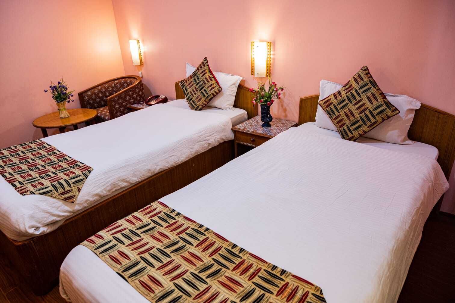 Quarto de hotel com duas camas individuais, cada uma com colchas e almofadas estampadas, uma cadeira de madeira, uma mesa pequena com um vaso de flores e luminárias fixadas na parede.