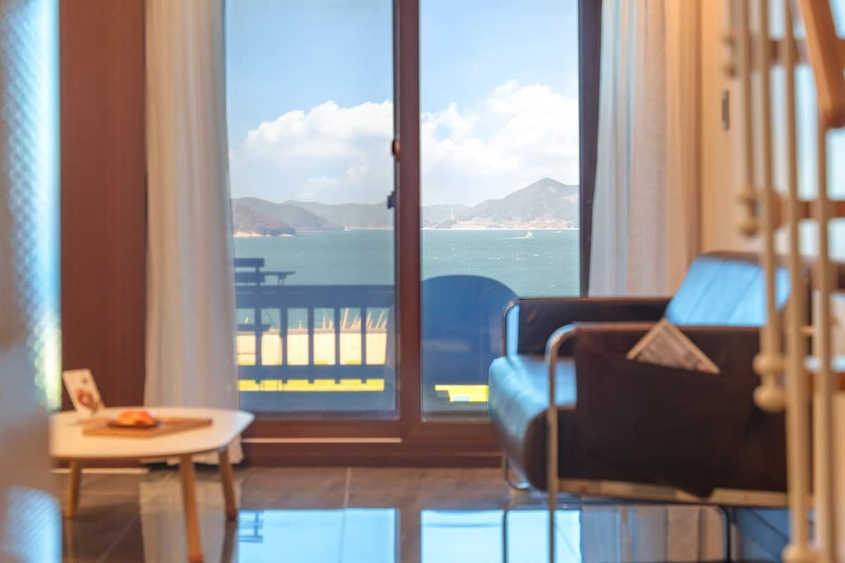 A room with a view of the sea through large glass doors, a sofa, and a small table with a plate.