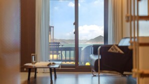 Habitació amb un gran finestral corredís que dona a un balcó amb vista al mar i muntanyes llunyanes.