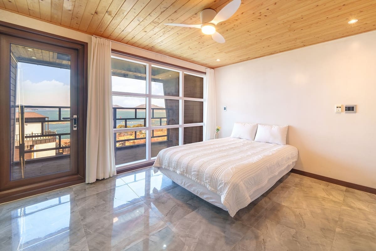 A bedroom with a large bed, a ceiling fan, and a view of the beach through a sliding glass door.