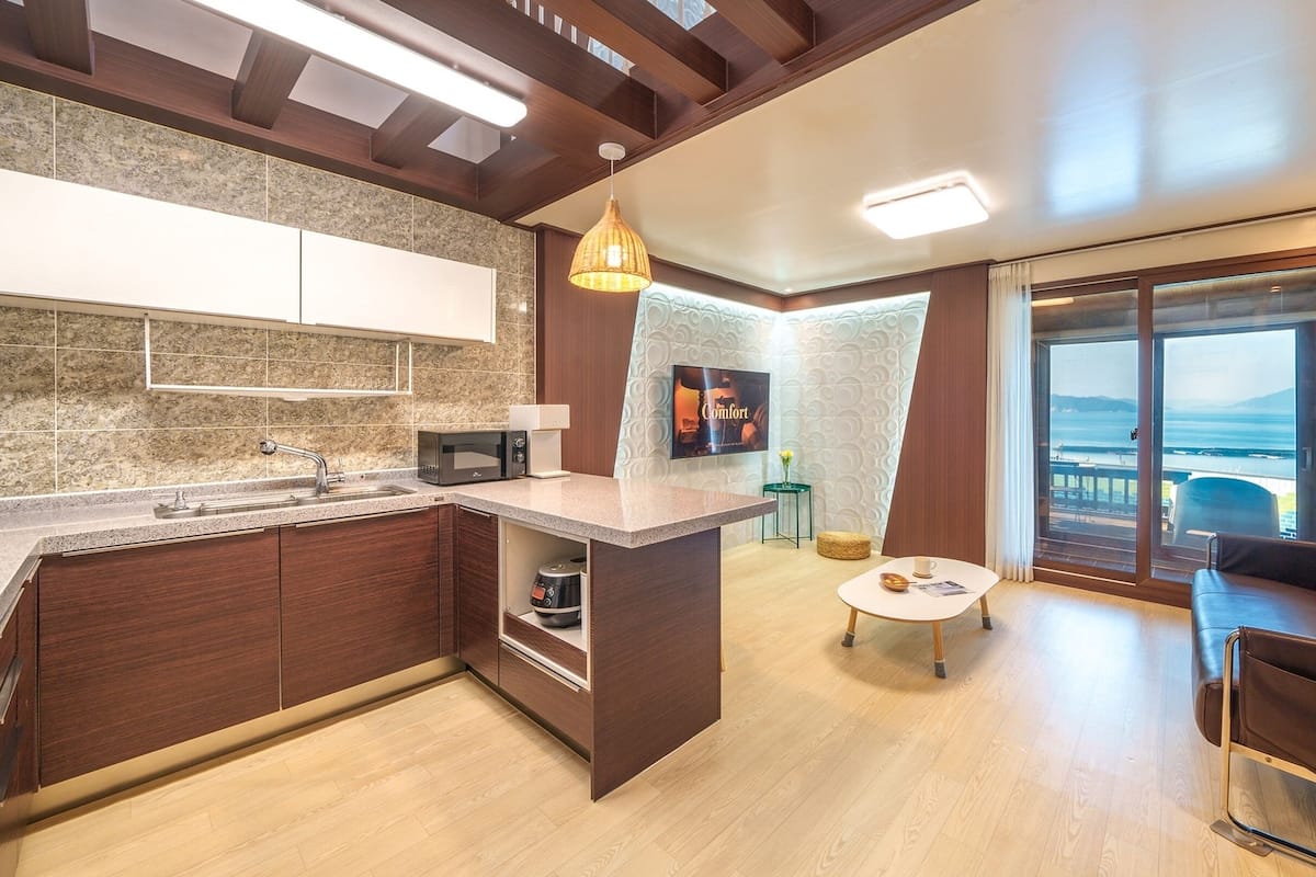 A modern kitchen with a built-in microwave, a sink, and a small island. There is a dining area with a table and chairs, and a large window offering a view of the sea.