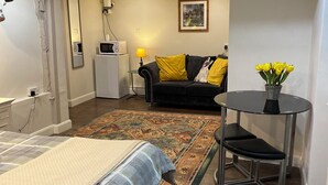 A bedroom with a bed, a small table with a vase of flowers, a sofa with yellow cushions, and a painting on the wall. - Old Thatch Bambers Green (Bishop's Stortford)