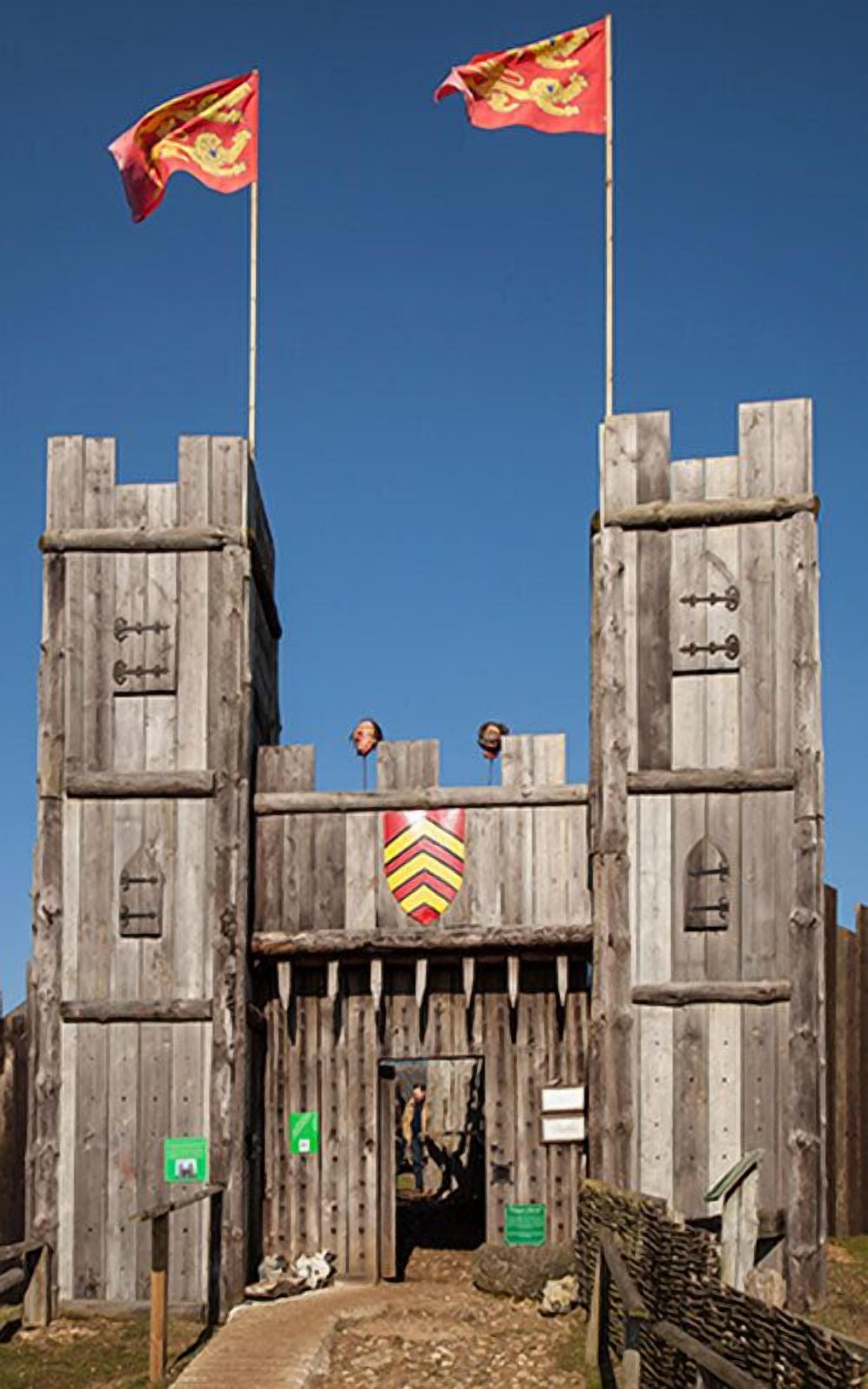 Entrada de un castillo de madera con dos banderas, un escudo y un letrero que indica un museo.