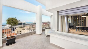 Ein Balkon mit Blick auf ein Kreuzfahrtschiff und den Hafen, weiße Wände sowie ein Essbereich mit schwarzen Stühlen und einem Tisch.
