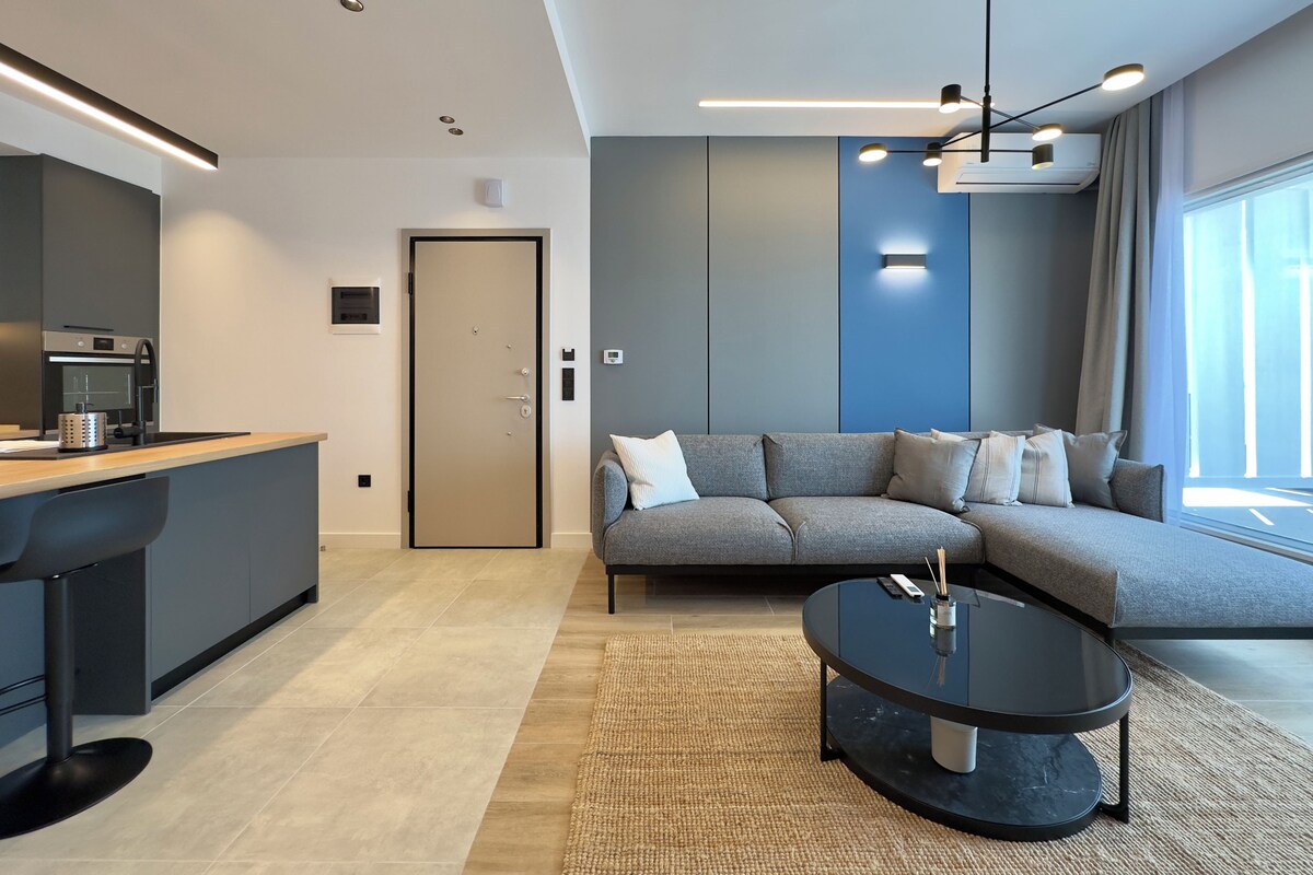 Una sala de estar moderna con un sofá gris, una mesa de centro negra y una zona de cocina con horno empotrado y fregadero.