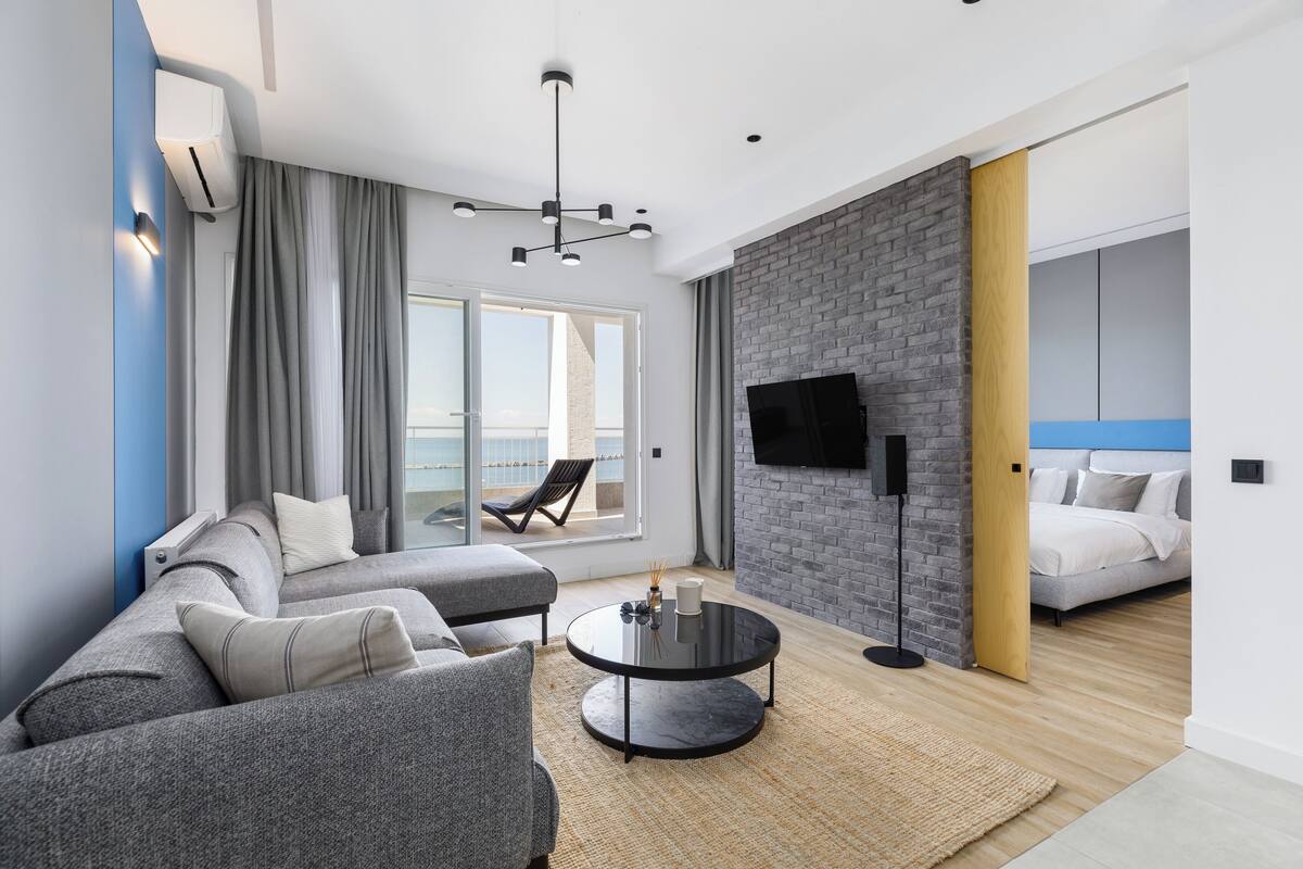 A modern hotel room with a grey sofa, a round coffee table, a flat-screen TV mounted on a brick wall, and a balcony with a chair.