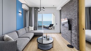 Ein modernes Wohnzimmer mit einem grauen Ecksofa, einem Glastisch und einem großen Fenster mit Meerblick.