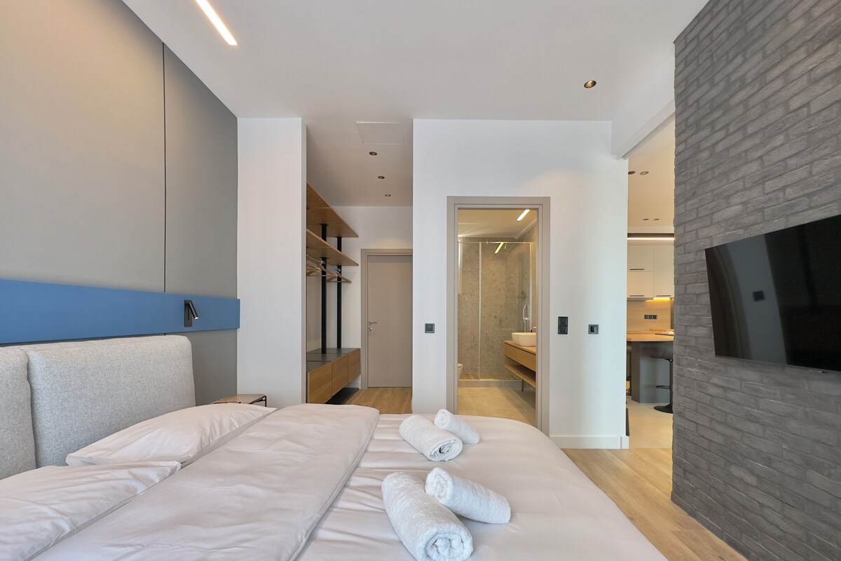 A modern hotel room with a large bed, a built-in wardrobe, a shower area, and a kitchenette.