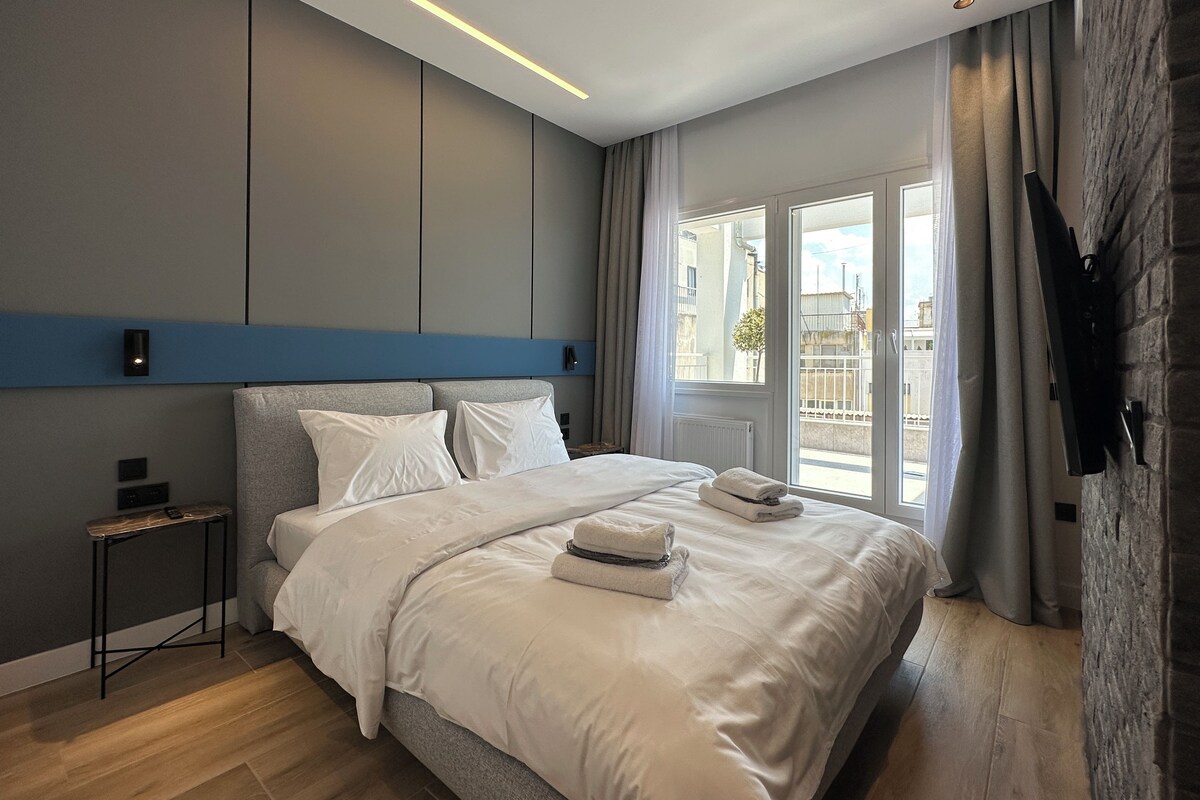 Una habitación de hotel moderna con una cama grande, una mesita de noche, un televisor de pantalla plana montado en la pared y vistas a la ciudad por la ventana.