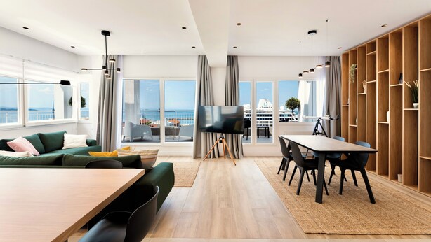 Ein modernes Wohnzimmer mit großem Fenster, einem Flachbildfernseher, einem Essbereich mit Tisch und Stühlen sowie einem Holzregal.