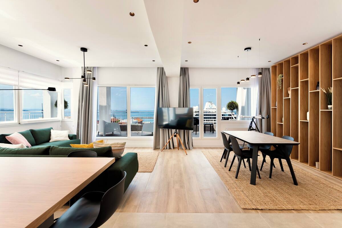 A modern living room with a large window, a flat-screen TV, a dining area with a table and chairs, and a wooden bookshelf.