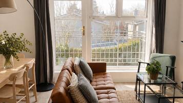 A modern living room with a leather sofa, a dining table with chairs, a glass coffee table, and a large window with curtains.