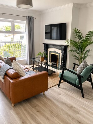 A modern living room with a leather sofa, a glass coffee table, a fireplace, and a TV mounted above it.