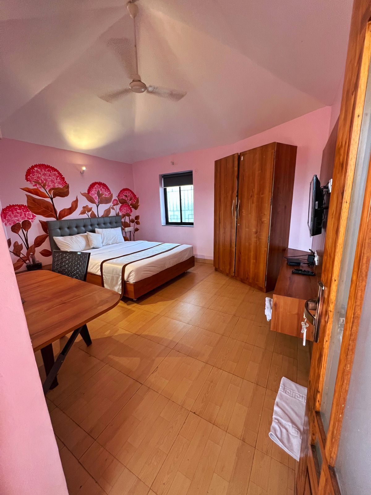 A bedroom with a bed, a wooden table, a ceiling fan, and a wardrobe.