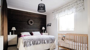 Een slaapkamer met een houten hoofdeinde, een bed met een grijze en witte deken, twee nachtkastjes met lampen, een ledikant, een raam met kantgordijnen en een wandkrans.