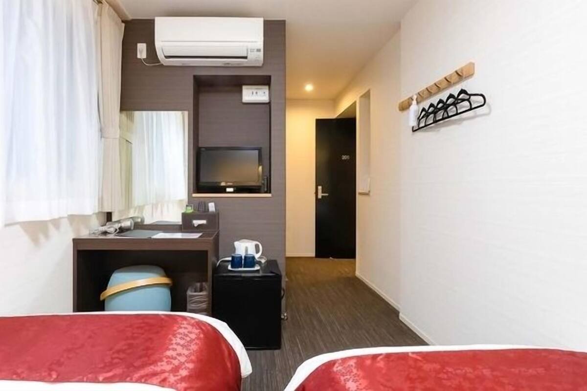 A hotel room with two beds, a desk with a computer, a TV, and an air conditioning unit.