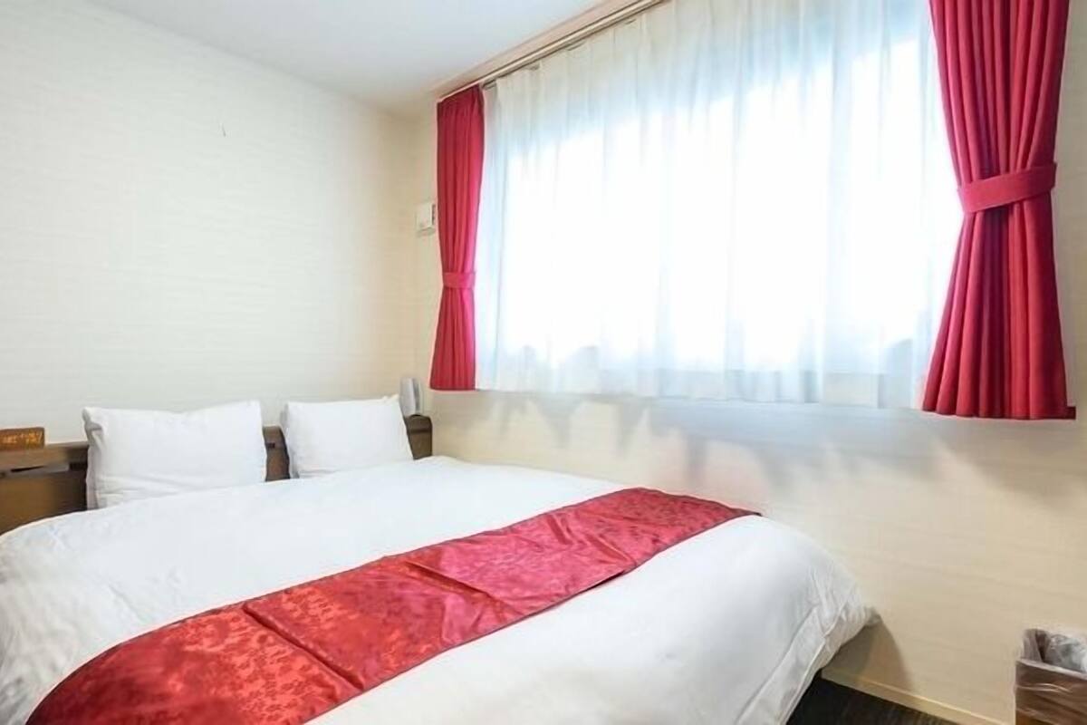 A hotel room with a large bed, white bedding, a red accent bedspread, a wooden nightstand, a red curtain, and a window with sheer white curtains.