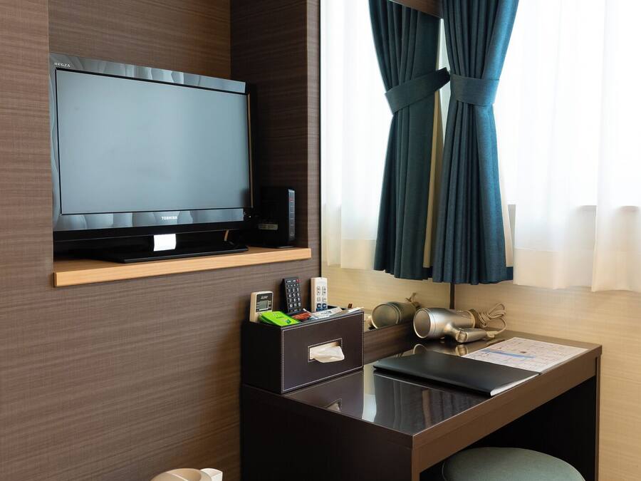 Een hotelkamer met een flatscreen-tv, een bureau met telefoon, een waterkoker en twee mokken, een klein voetenbankje en een raam met gordijnen.
