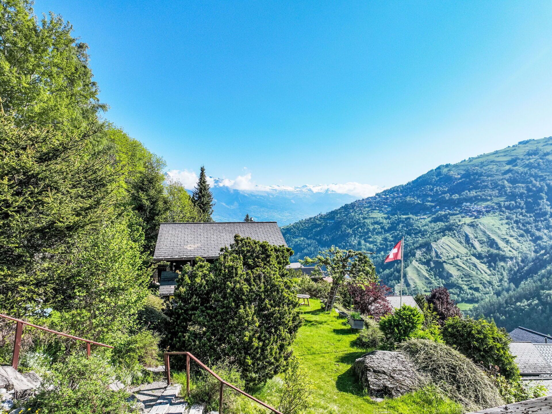 Uma cabana de montanha com uma bandeira suíça, rodeada por vegetação exuberante e uma vista clara de montanhas distantes.
