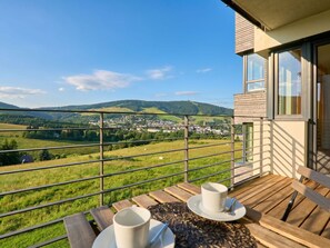 Un balcon avec une table et des chaises, donnant sur un paysage pittoresque composé de collines et d’une ville.