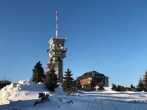 Una torre alta con múltiples antenas, rodeada de nieve y árboles, con un edificio al fondo.