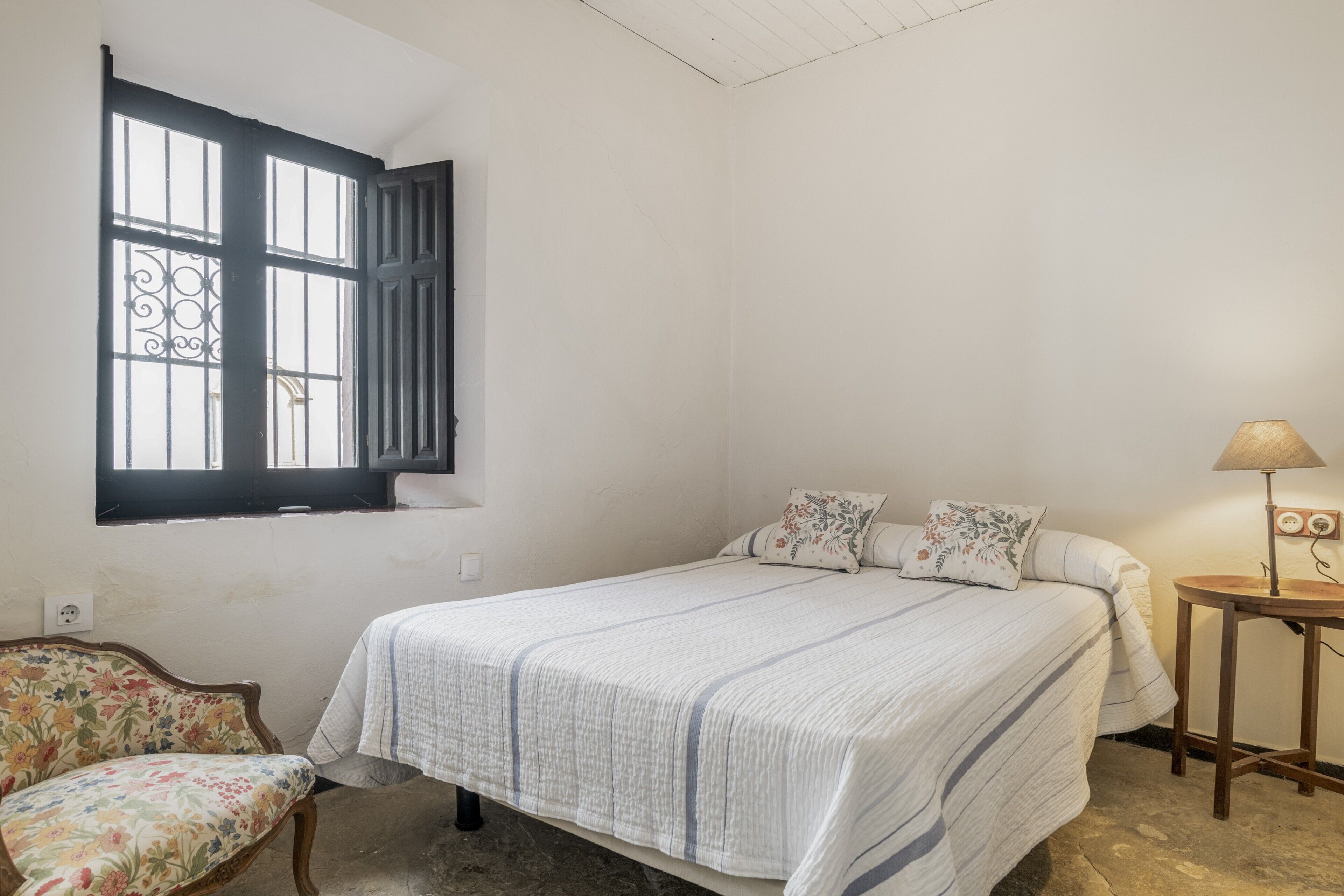 A bedroom with a bed, a chair, a window with bars, and a lamp.