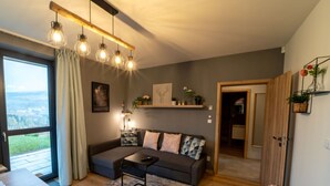 A modern living room with a grey sofa, a small table, and a wooden floor. There is a large window with curtains, a ceiling light fixture, and a door leading to another room. - Cosy apartment for 4 guests with WIFI, TV and terrace (Klášterec Nad Ohrí)