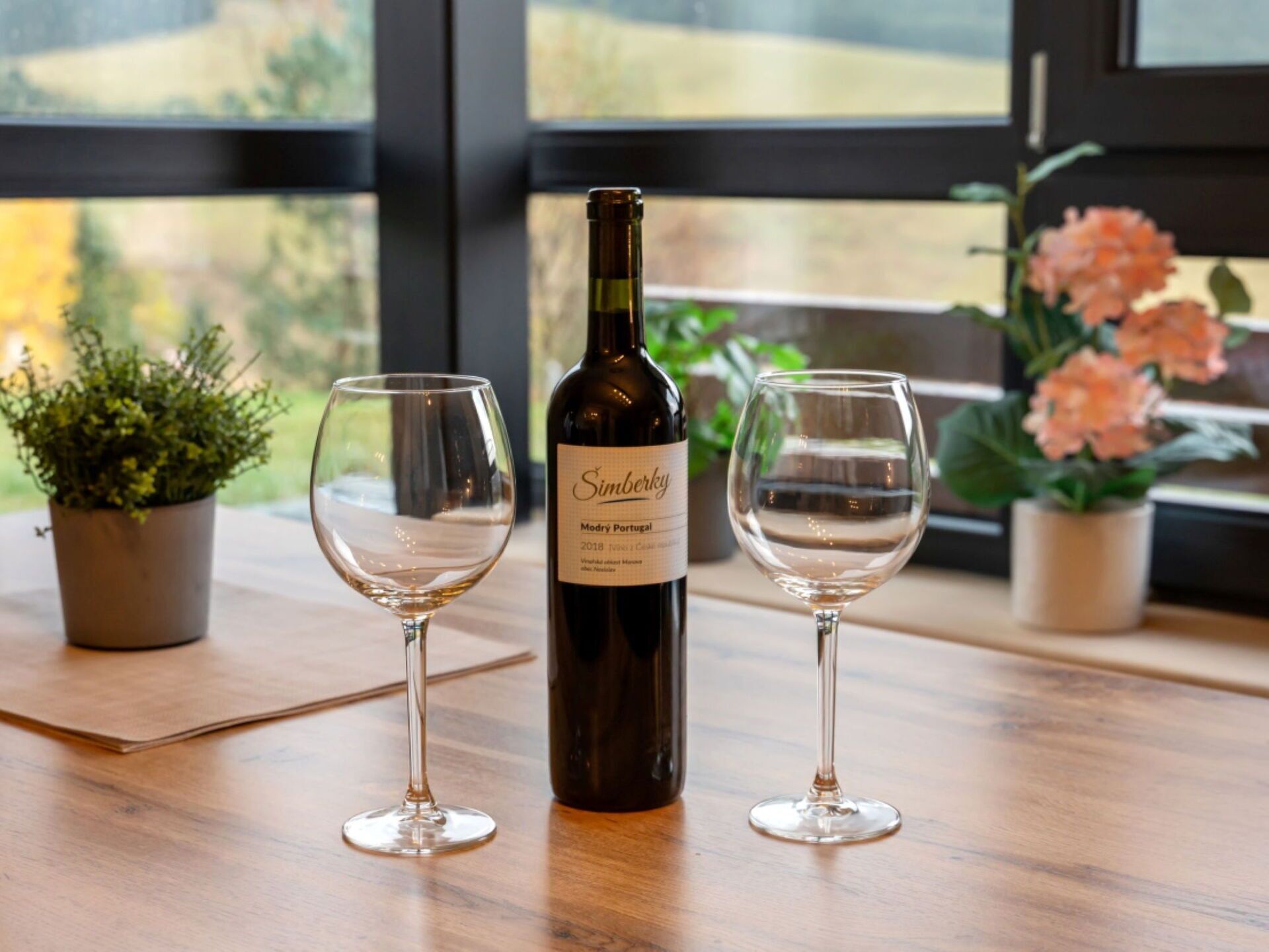 Une bouteille de vin et deux verres sur une table en bois près d’une fenêtre ornée de plantes.