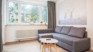 A modern living room with a grey sofa, a small white table, and a radiator. There is a window with curtains and a view of trees outside. - Beautiful apartment for 4 guests with WIFI, TV and balcony (Winterberg)
