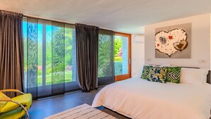 Ein Schlafzimmer mit einem großen Bett, einem grünen Sessel, Blick ins Grüne durch das Fenster und einem Wandbild.