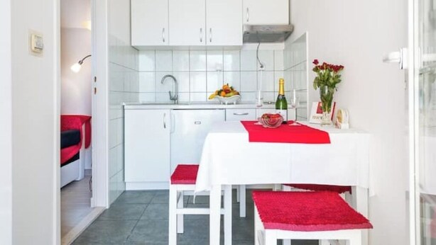A compact kitchen with white cabinets, a white countertop, and a dining table with red accents.