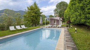 Ein Schwimmbecken mit Liegestühlen und einer überdachten Sitzgelegenheit, umgeben von üppiger Vegetation und Bergen im Hintergrund.