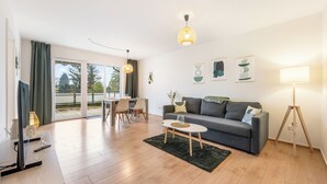 A modern living room with a grey sofa, a small coffee table, and a TV on a stand. There is a dining area with a table and chairs, and a balcony with a view of trees. - Cozy barrier-free apartment in central Vegesack/2-4 pers. (Bremen)