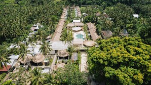 An aerial view of a resort with multiple thatched-roof structures, a central swimming pool, and lush tropical vegetation. - Lombok Souls (Senggigi)