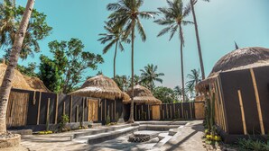 A tropical resort with thatched-roof huts, palm trees, and a stone pathway leading to a central seating area. - Lombok Souls (Senggigi)