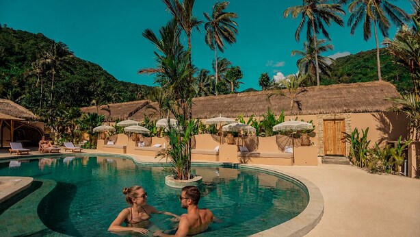 A pool area with lounge chairs, palm trees, and thatched-roof structures. - Lombok Souls (Senggigi)