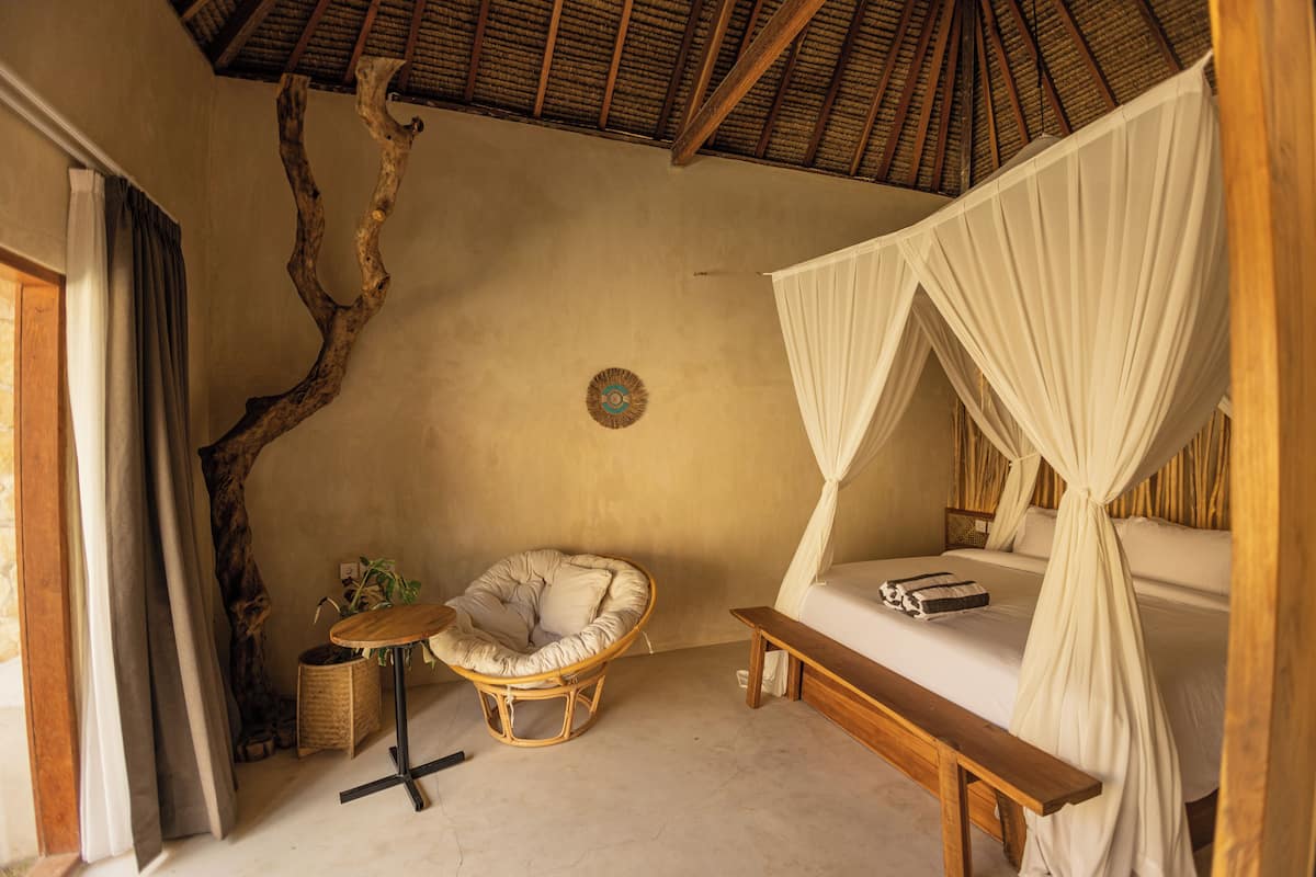 A room with a bed, a chair, a small table, and a large tree branch against the wall.