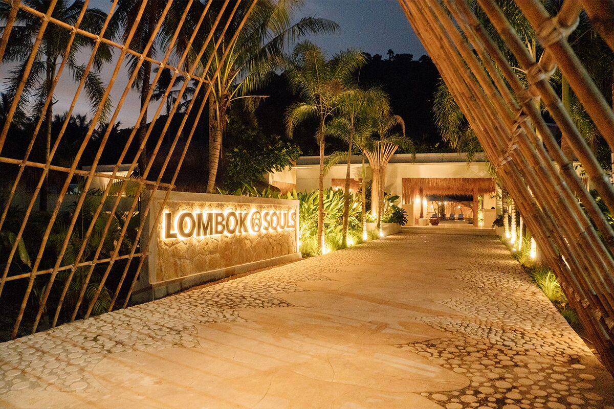 Ein beleuchteter Eingang zu einer Unterkunft namens 'Lombok Souls' mit einem Steinweg, Bambuszaun und Palmen.